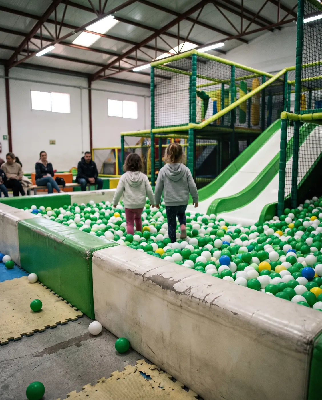Dos niños de espaldas cruzan una piscina de bolas verdes y blancas hacia un tobogán (foto ilustrativa generada con IA)
